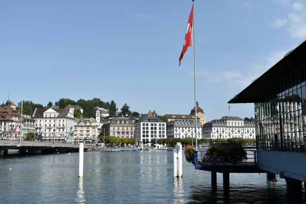 Blick auf eine Schweizer Stadt am Wasser mit einer wehenden Schweizer Flagge bei klarem Himmel – symbolisch für die Ankunft in der Schweiz.